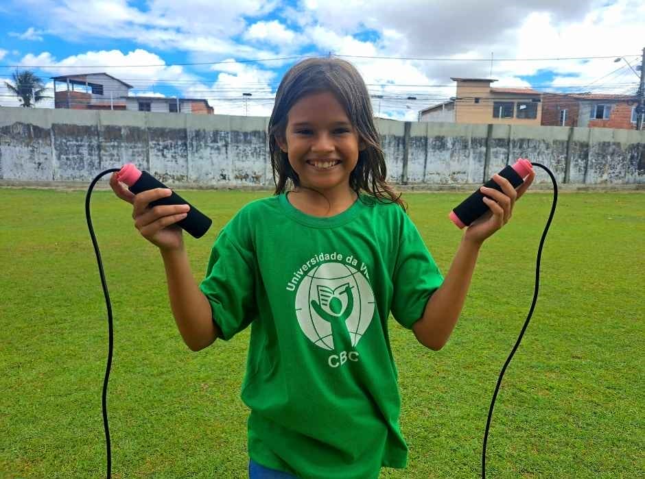 Girl with jump rope standing in front of green field and wall with favelas behind it